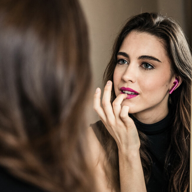 Frau mit lila kabellosen Bluetooth Kopfhörern steht vor einem Spiegel und trägt lila Lippenstift auf