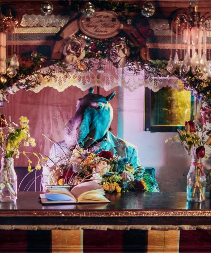 Verkleidete Person in Nilpferd Maske sitzt an einer bunt dekorierten Theke